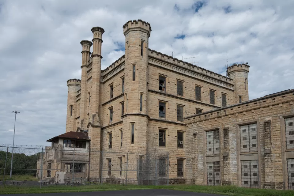 Deralict stone prison building with turrets and a chain link fence in foreground, under a gray, cloudy sky.